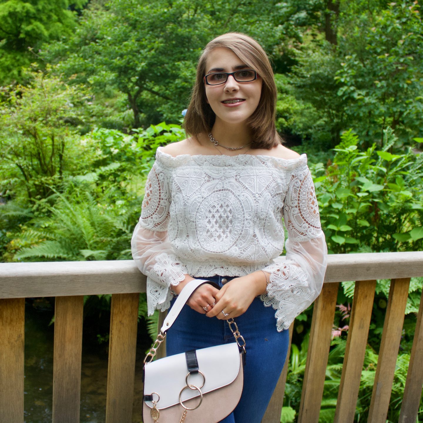 A photo of Elin leaning on a wooden bridge, she is smiling at the camera and she wears a white bardot top, blue skinny jeans and a white and beige saddle bag