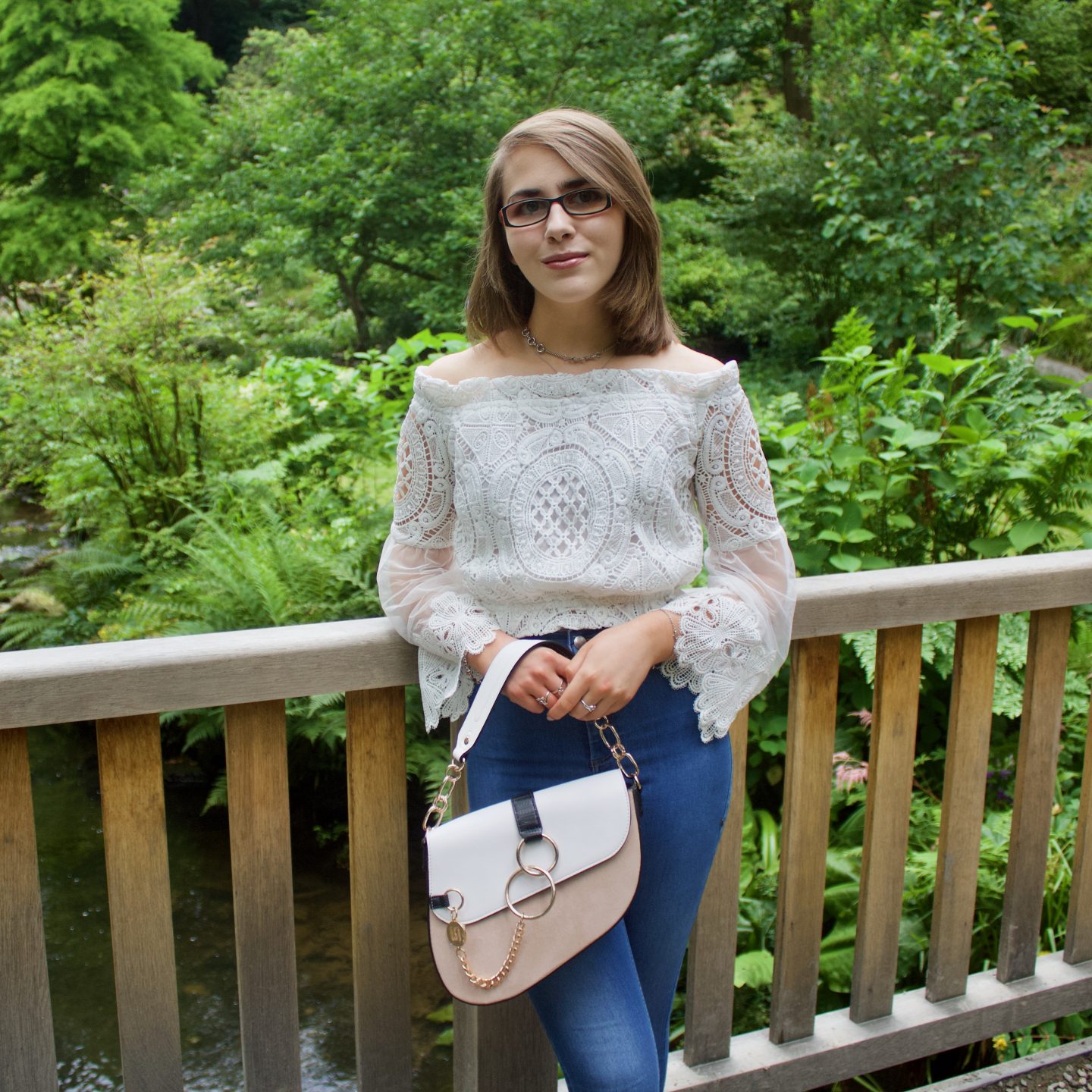 A photo of Elin leaning against a wooden bridge, she is holding a white and beige saddle bag which has gold hardware and there are green plants and trees in the background