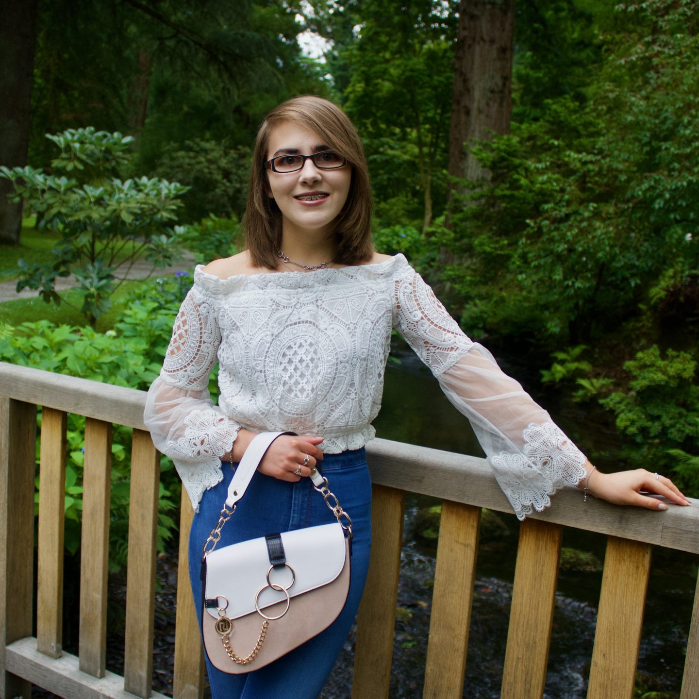 A photo of Elin leaning against a wooden bridge, there are trees in the background and she is smiling at the camera