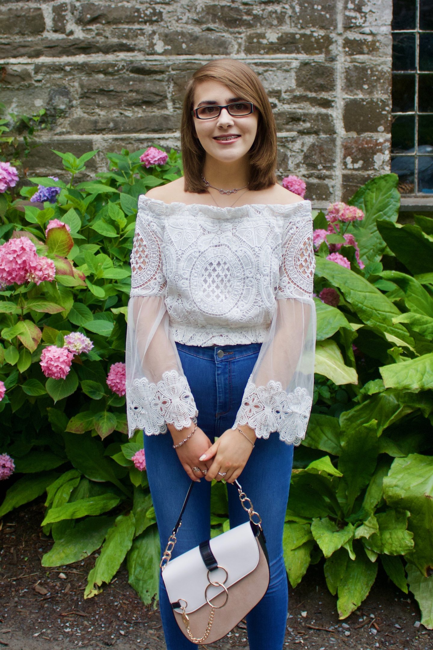 A photo of Elin wearing blue skinny jeans and a white lace bardot top which has fluted sleeves, she is also holding a white and beige saddle bag and she stands in front of a pretty stone wall and pink flowers