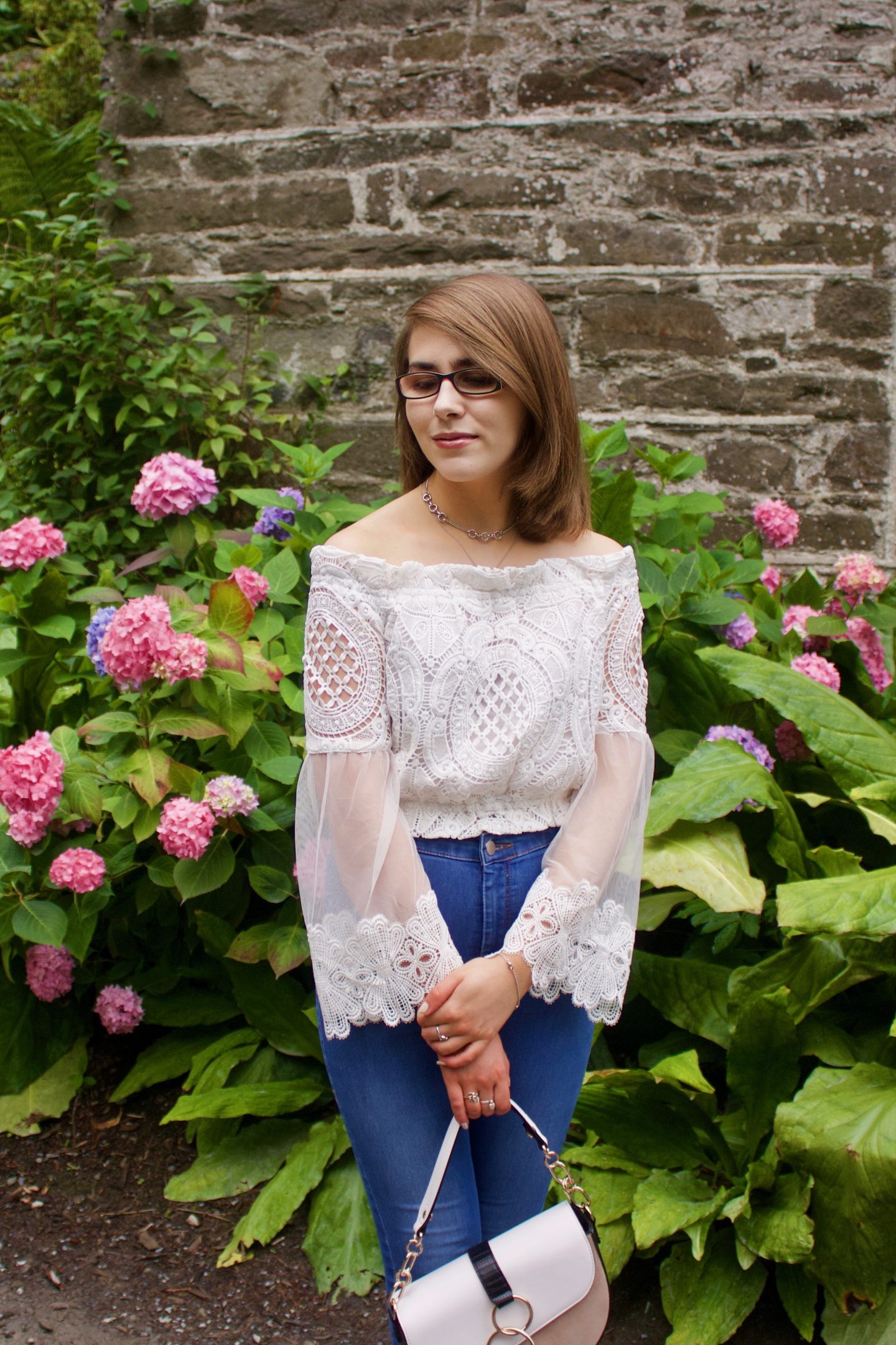 A photo of Elin wearing blue skinny jeans and a white lace bardot top which has fluted sleeves, she is also holding a white and beige saddle bag. She is looking down and she stands in front of a pretty stone wall and pink flowers