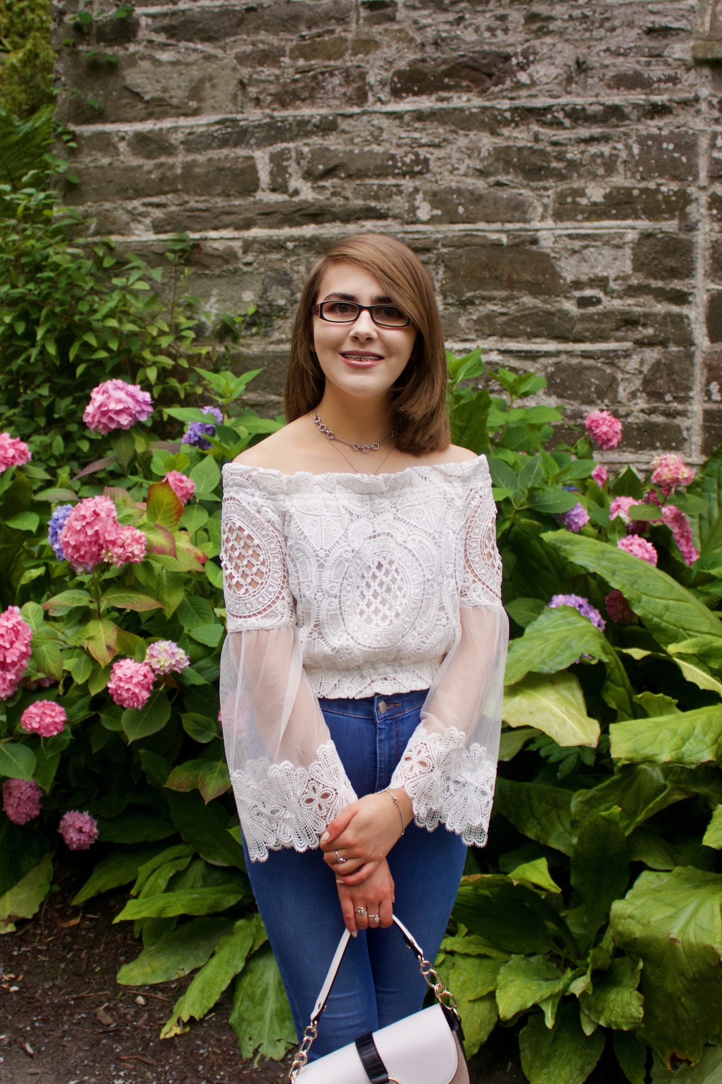 A photo of Elin wearing blue skinny jeans and a white lace bardot top which has fluted sleeves, she is also holding a white and beige saddle bag and she stands in front of a pretty stone wall and pink flowers