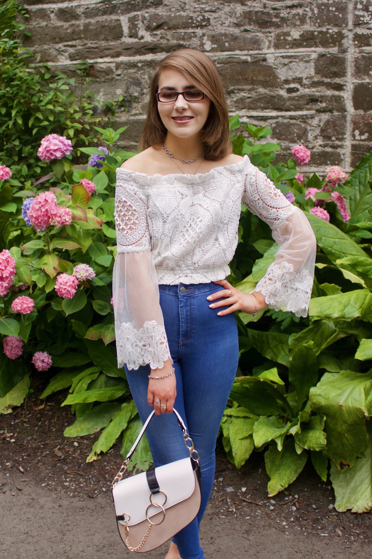 A photo of Elin wearing a white lace mesh bardot top, blue skinny jeans, white slip on shoes and a white and beige saddle bag, she is smiling and looking down and her left hand is on her hip. There is a stone wall and pink flowers in the background