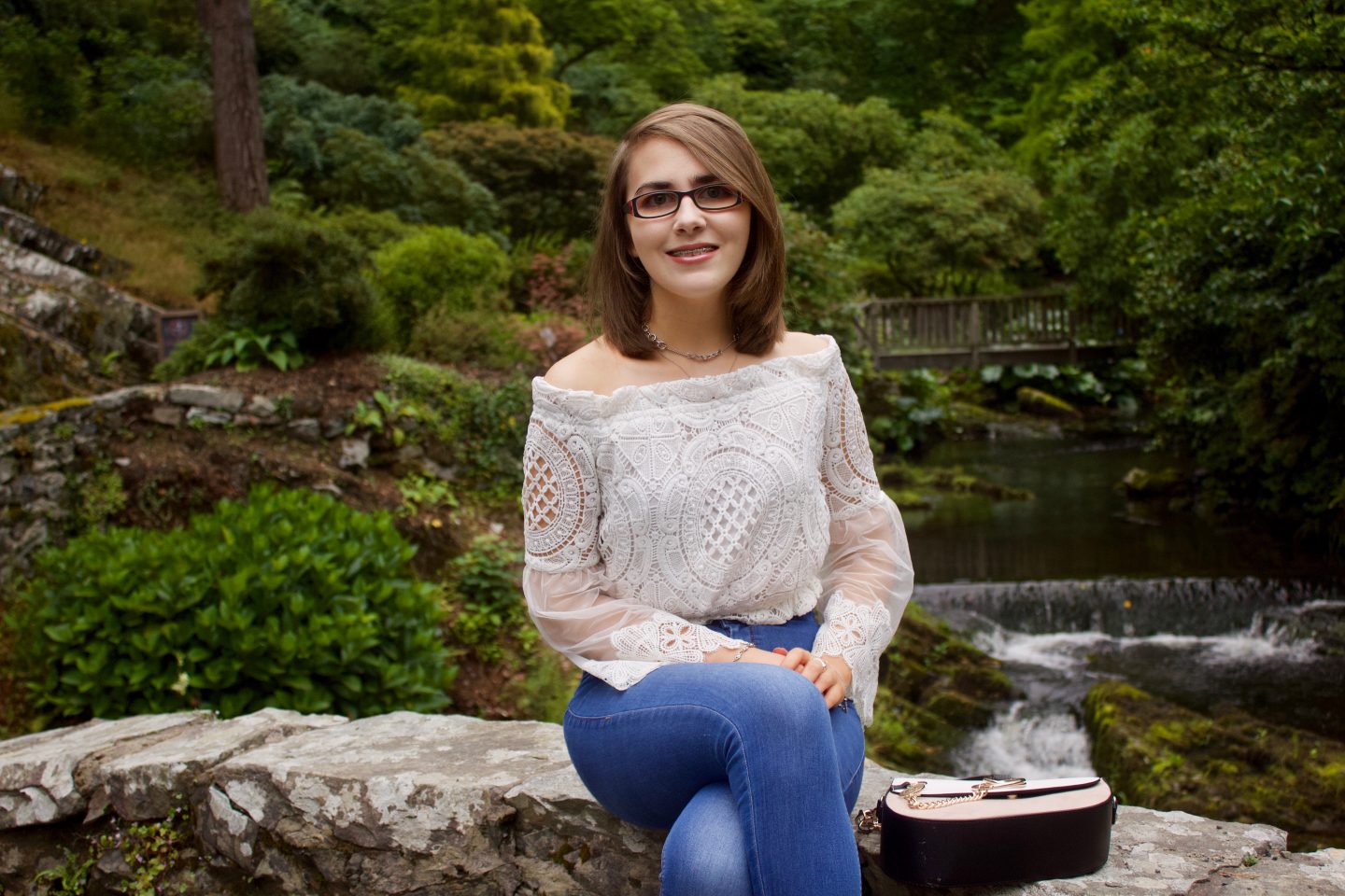 A photo of Elin sitting on a small stone bridge, she is wearing a white lace bardot top, blue skinny jeans and two silver necklaces, there are trees and flowers in the background