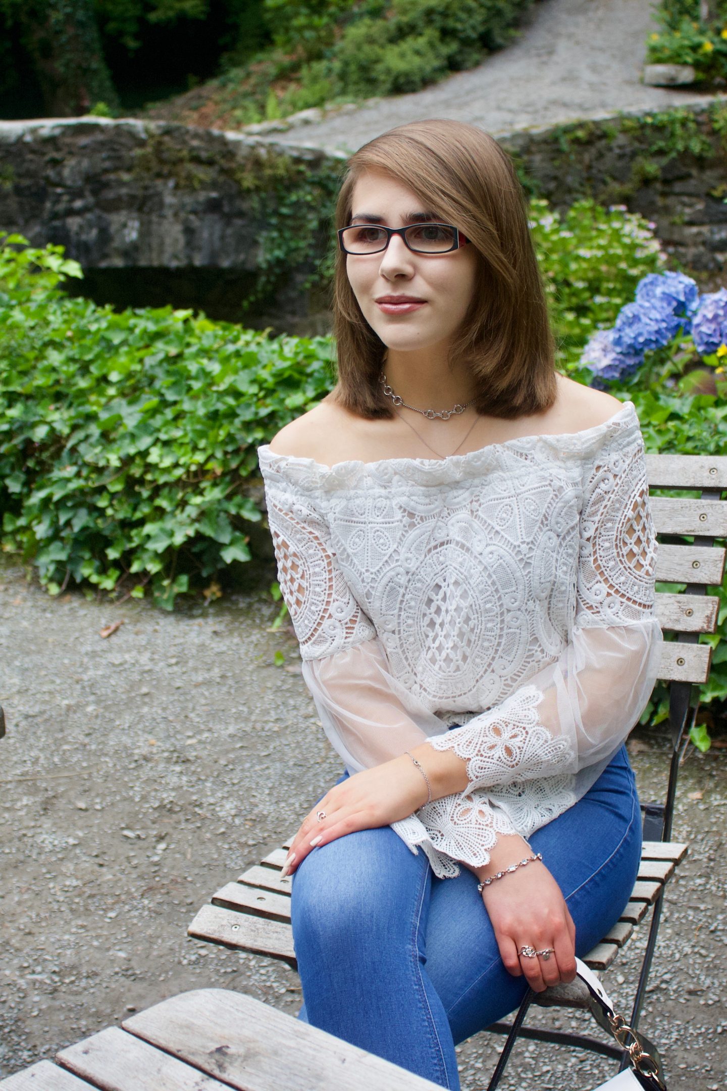 A photo of Elin sitting on a wooden chair in front of a small stone bridge, she is wearing a white lace mesh top and blue skinny jeans and is looking away from the camera