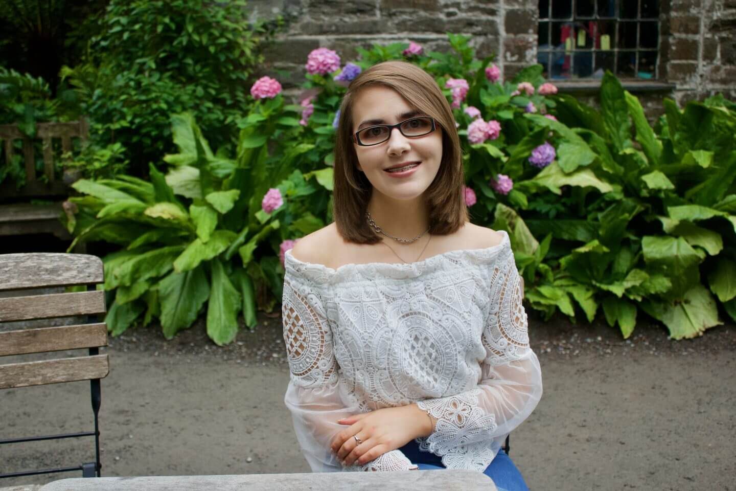 A photo of Elin sitting on a wooden chair in front of a pink flowers and a pretty stone building, she is wearing a white lace mesh bardot top and is smiling at the camera