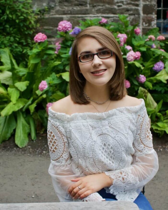 A photo of Elin sitting on a wooden chair in front of a pink flowers and a pretty stone building, she is wearing a white lace mesh bardot top and is smiling at the camera