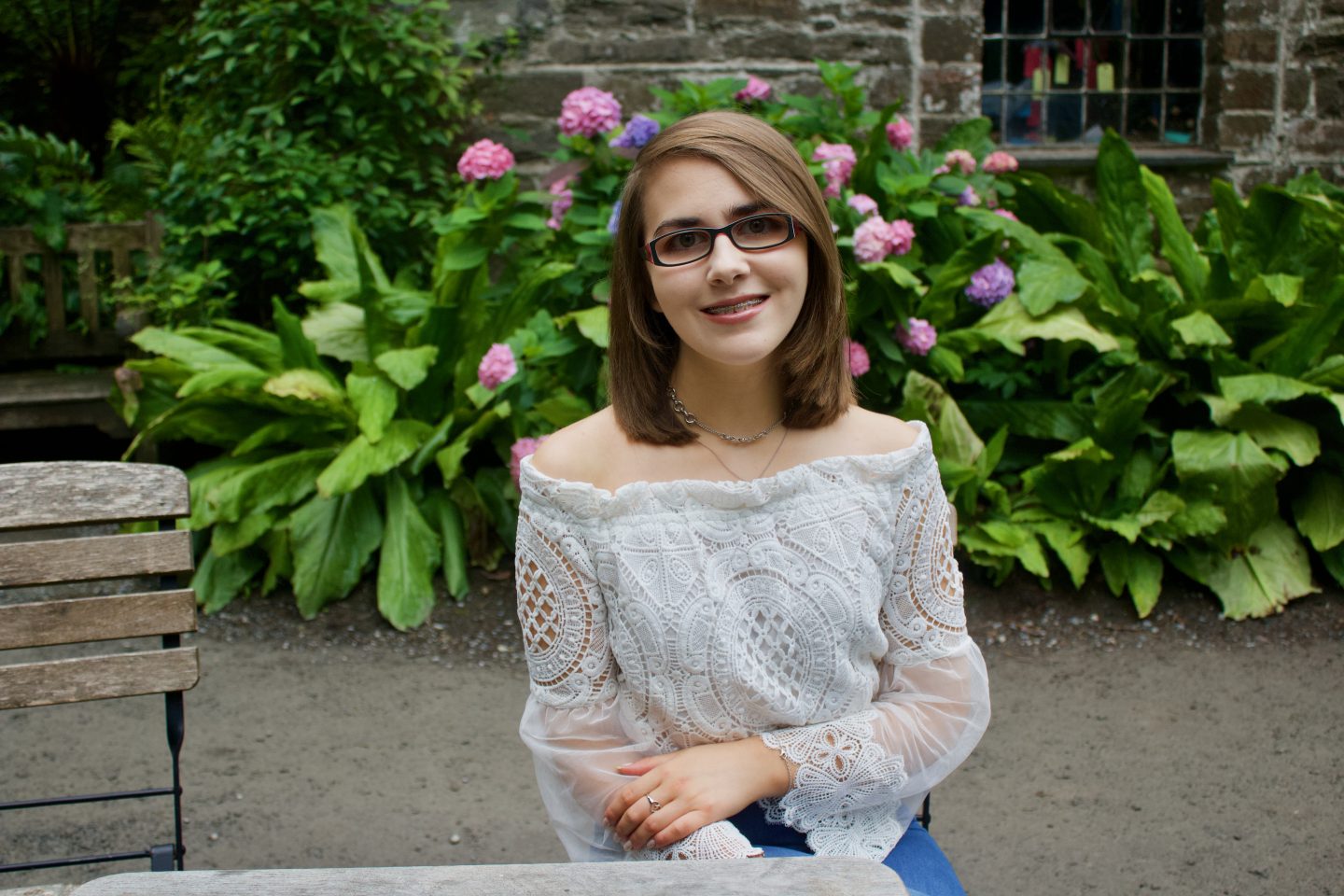 A photo of Elin sitting on a wooden chair in front of pink flowers and a pretty stone building, she is wearing a white lace mesh bardot top and is smiling at the camera