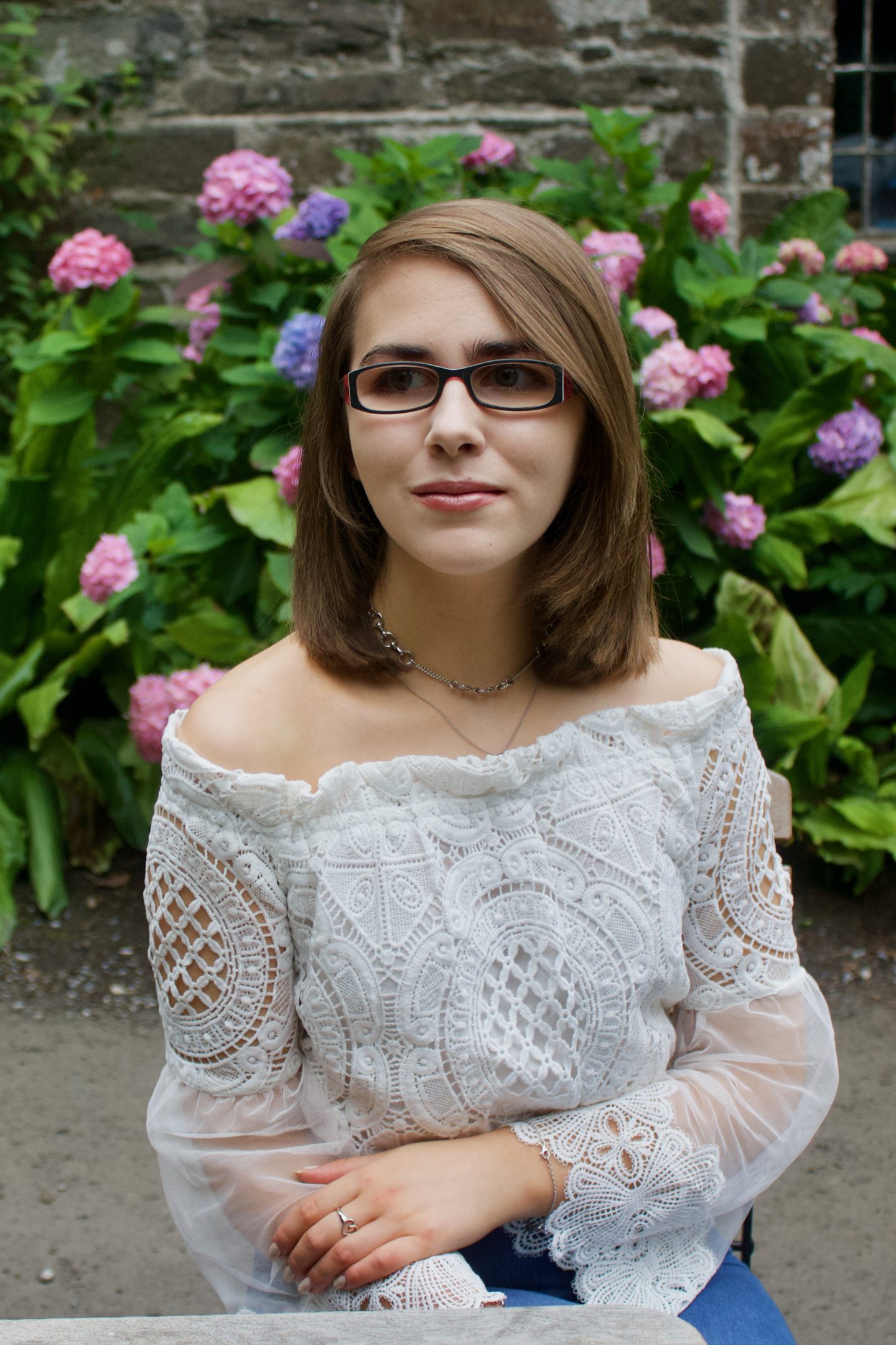 A photo of Elin sitting on a wooden chair in front of a pink flowers and a pretty stone building, she is wearing a white bardot top and is looking away from the camera