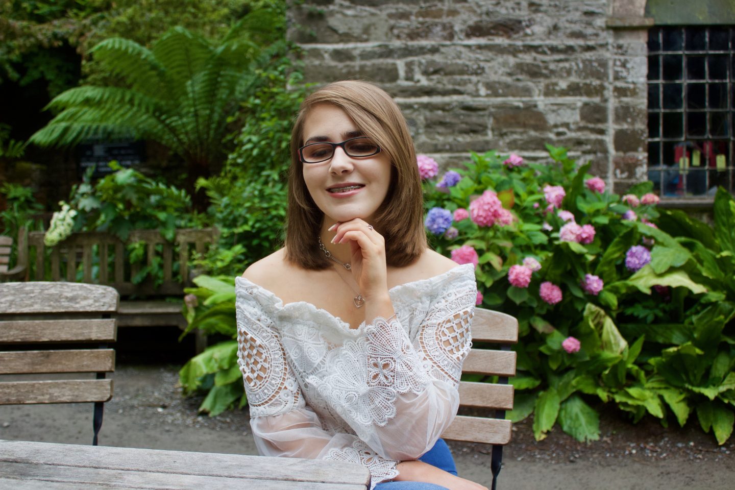 A photo of Elin sitting by a wooden table, she is looking down and smiling and her left hand rests underneath her chin. There is a stone building and pretty pink flowers in the background