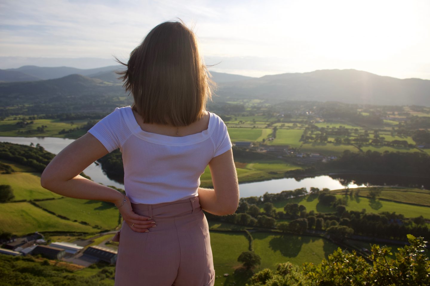 A photo of Elin from the back, one of her hands is on her hip and she is looking out at a view of mountains, fields and a river