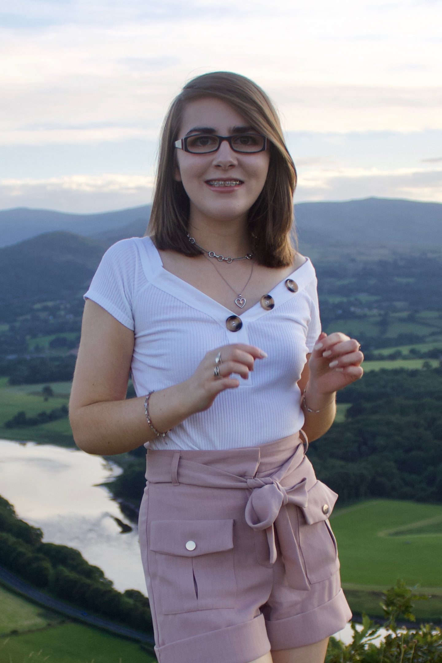 A photo of Elin wearing a white bardot top which has buttons on the neckline and high waisted pink utility shorts - she is smiling at the camera
