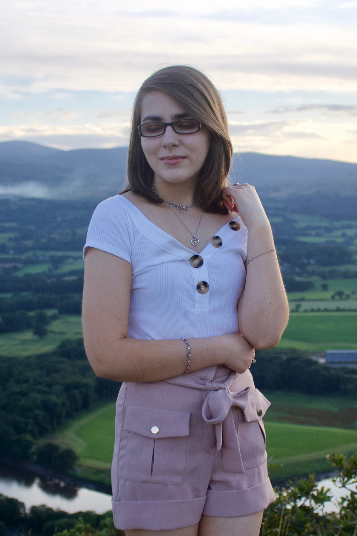 The fear of missing out as a blind person in a visual world: A photo of Elin wearing a white bardot top which has buttons on the neckline and high waisted pink utility shorts - she is looking down and her hand is raised, holding a strand of her hair