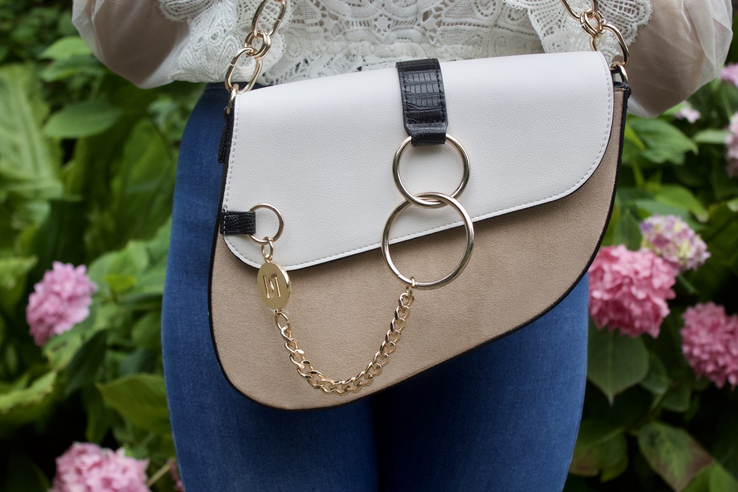 A photo of a white and beige saddle bag being held up with pink flowers to be seen in the background