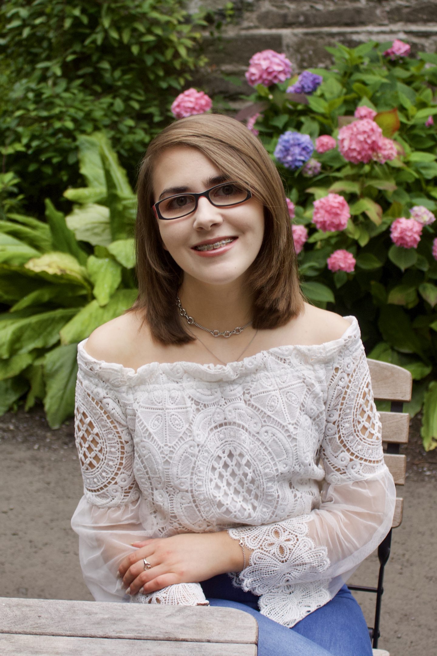 A photo of Elin sitting on a wooden chair in front of a pink flowers and a pretty stone building, she is wearing a white lace mesh bardot top and is smiling at the camera