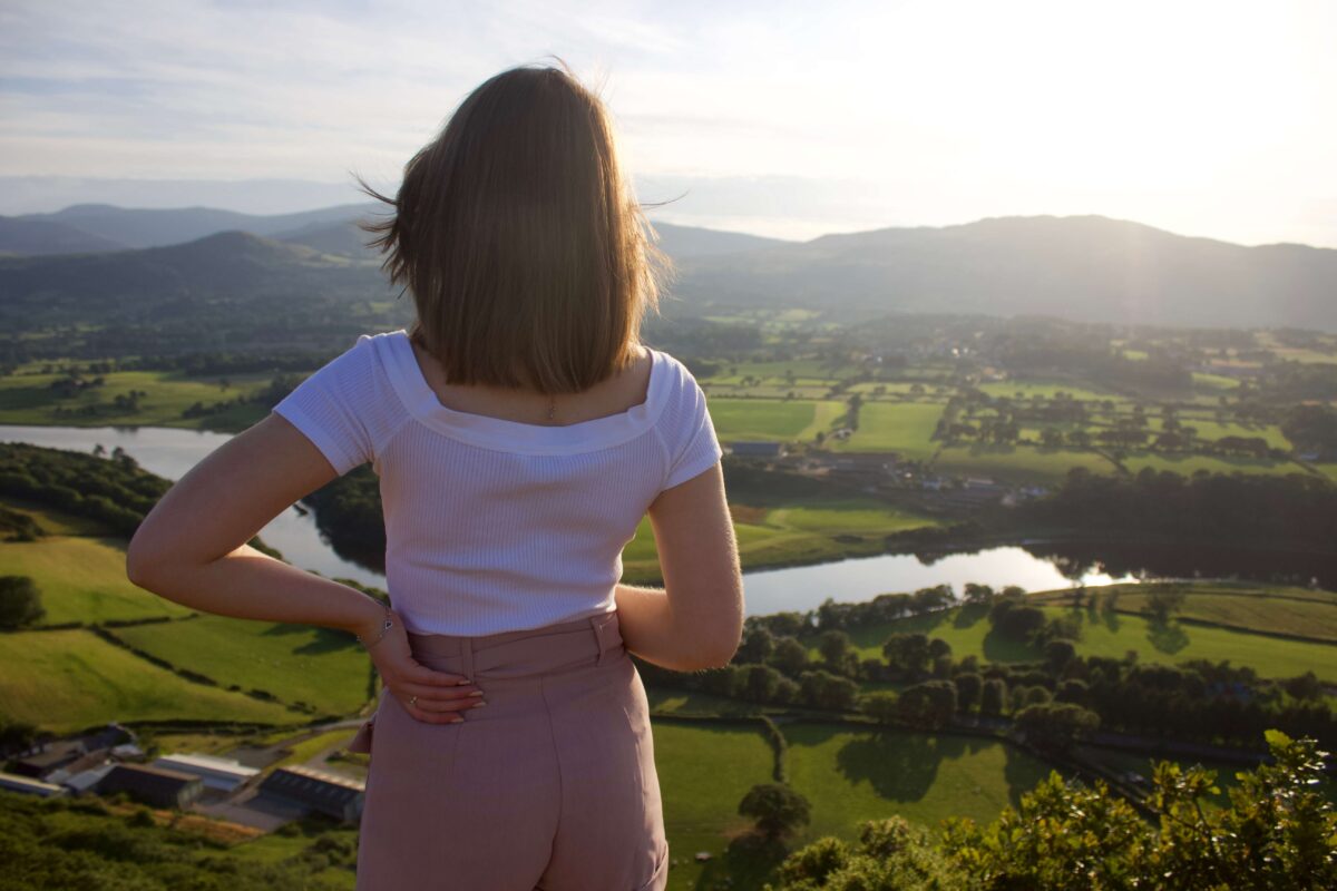 A photo of Elin from the back, one of her hands is on her hip and she is looking out at a view of mountains, fields and a river