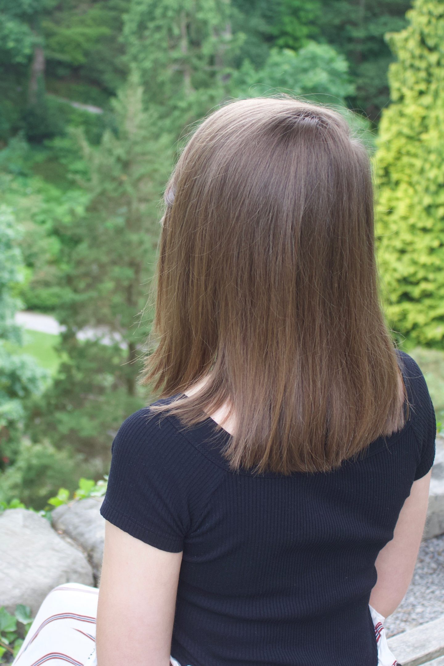 A photo of Elin from the back, she is sitting on a wooden bench and looking out on trees and flowers