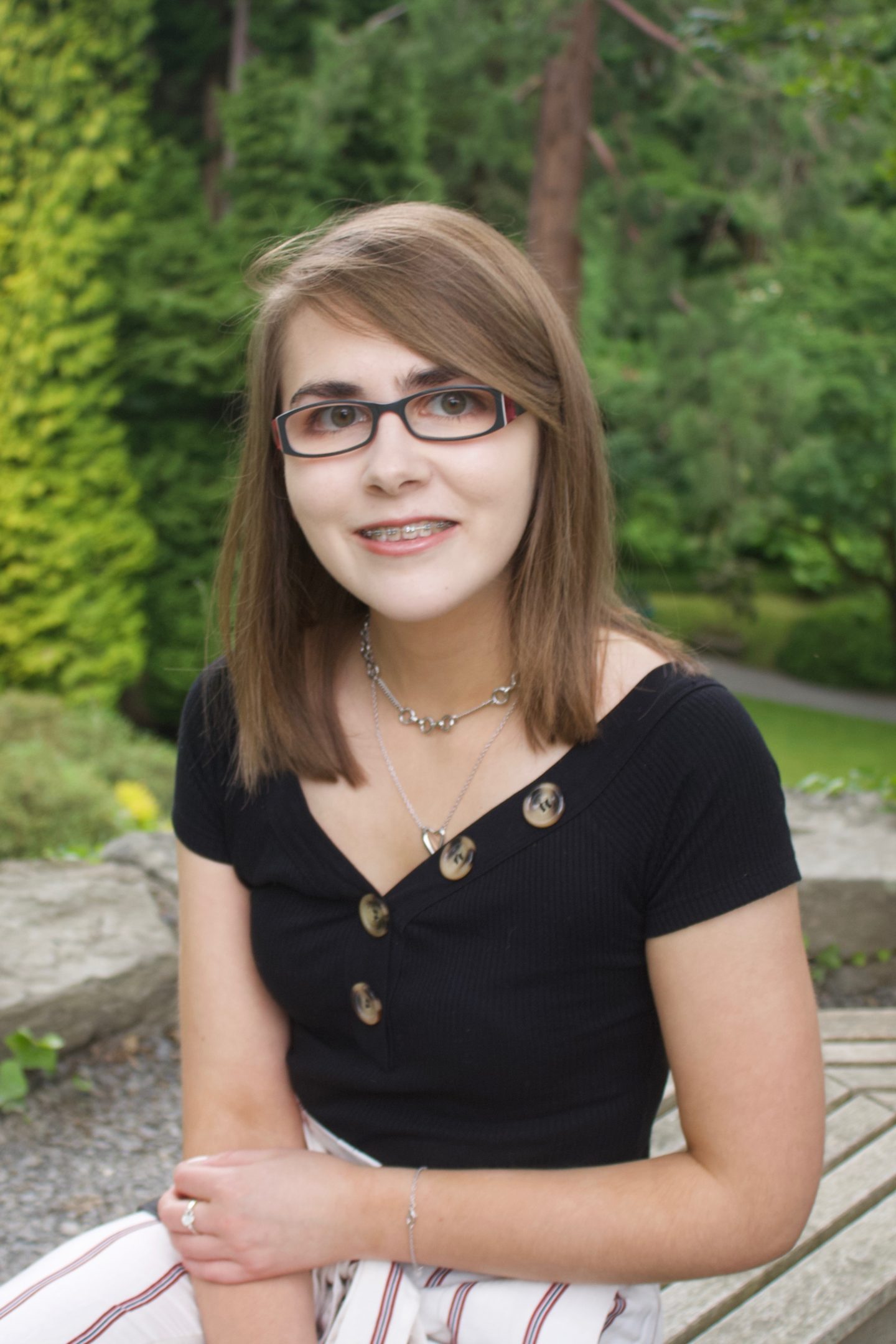 A photo of Elin sitting on a wooden bench which circles around a tree, there are trees in the background