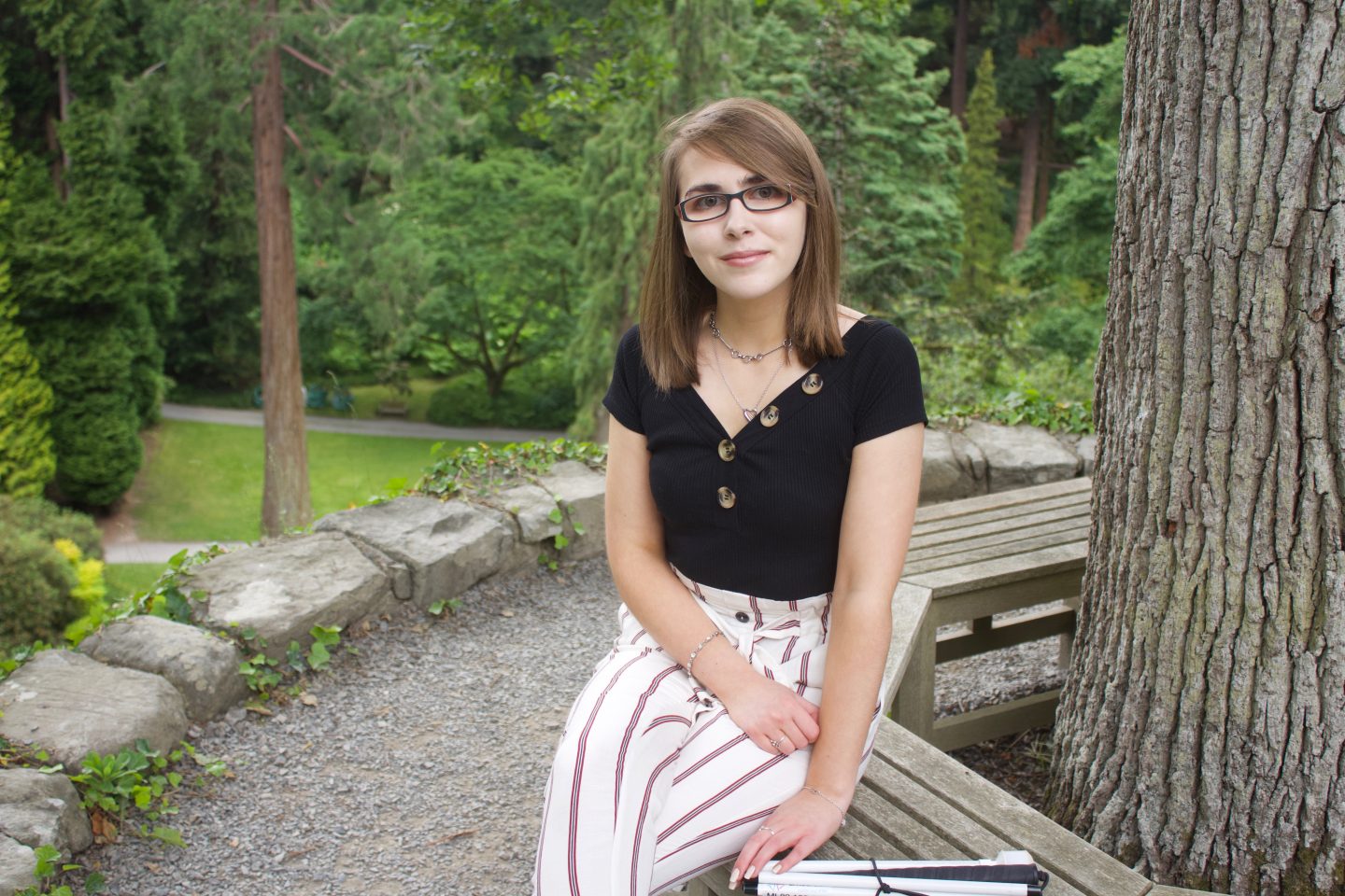 A photo of Elin sitting on a bench which circles around a tree, she wears cream trousers with red striped and a black bardot top which has buttons on the neckline