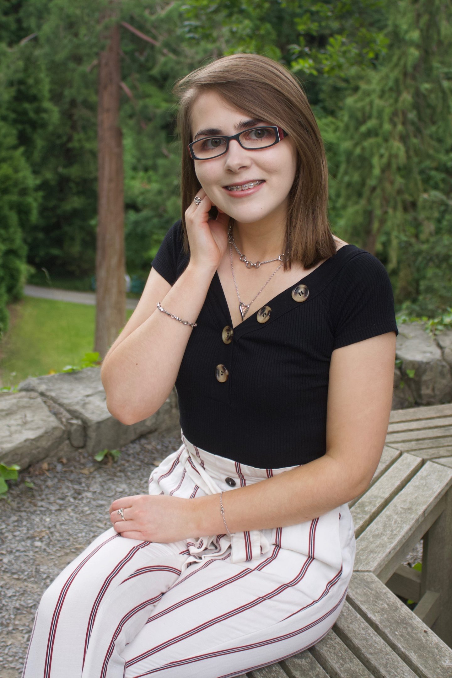 A photo of Elin sitting on a wooden bench with trees in the background. She is wearing a pair of cream skinny trousers which have red stripes and a black bardot top which has buttons on the neckline. One of her hands is raised to just under her ear.