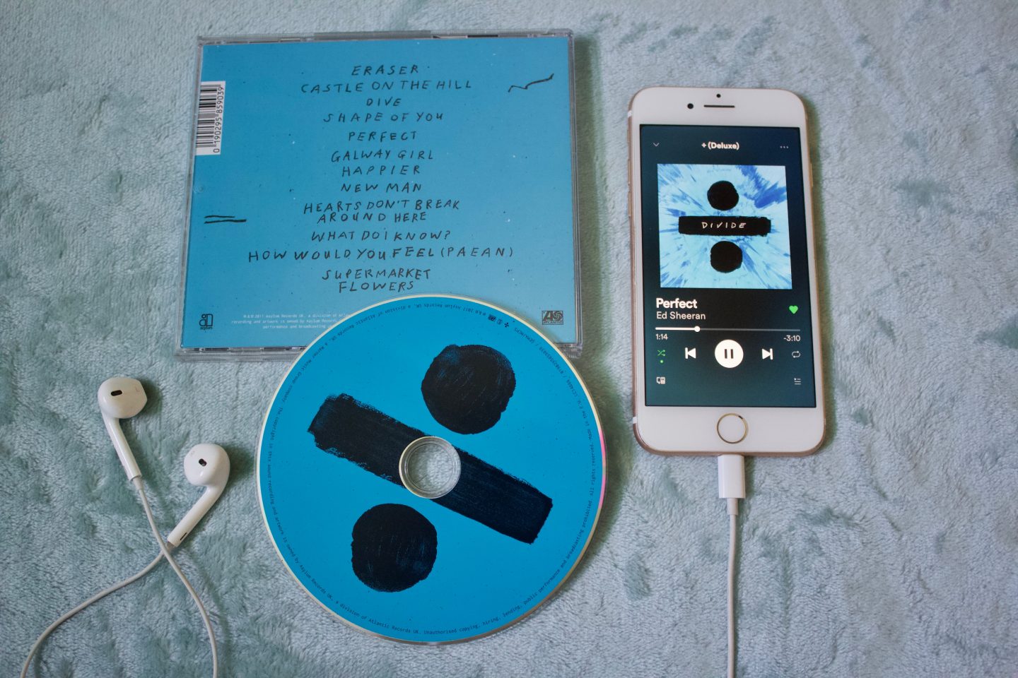 A photo of the back of the Divide album which shows the names of the songs, the CD itself is placed halfway on the case, my phone is beside it along with some earphones