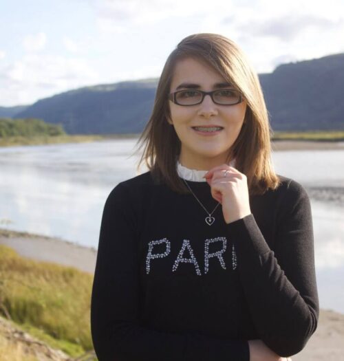 A photo of Elin standing in front of a river and mountains, she's smiling at the camera and her hand is raised to just underneath her chin