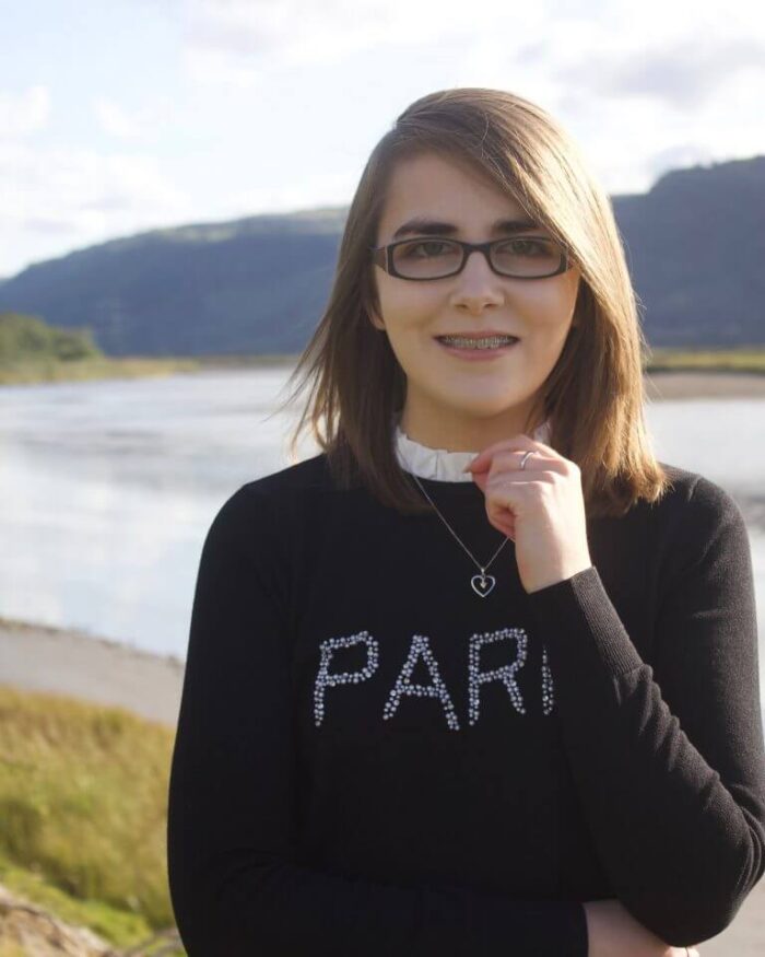 A photo of Elin standing in front of a river and mountains, she's smiling at the camera and her hand is raised to just underneath her chin