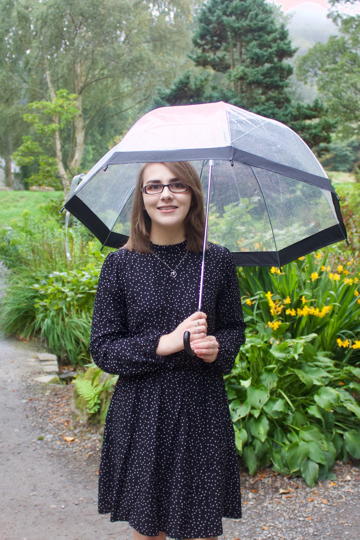 A photo of Elin standing in front some yellow flowers, she is holding up a clear umbrella and she's smiling at the camera
