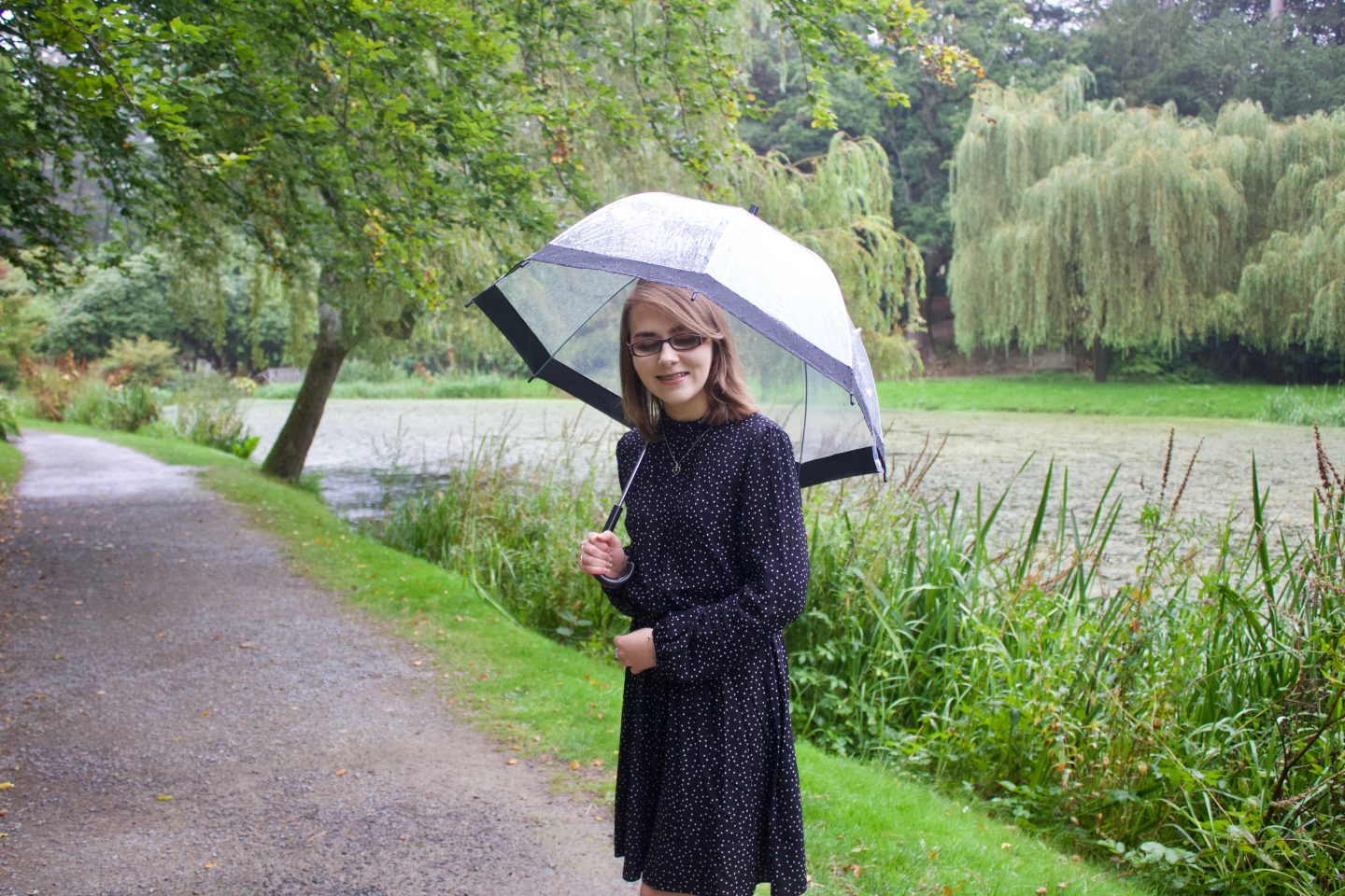 A photo of Elin standing in front of a small lake which has trees and flowers surrounding it. She is wearing a black skater dress which has small white polka dots on it and she's holding up a clear umbrella