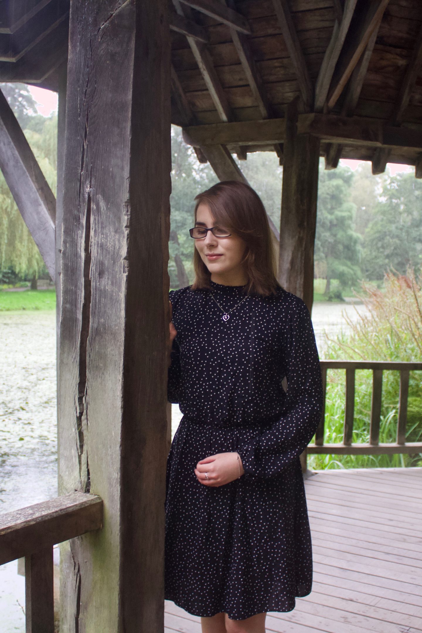 A photo of Elin leaning against a wooden pillar in the small boat house, she is looking down and wears a black skater dress which has white polka dots. She has accessorised with a small heart shaped necklace and a few silver rings, one of them matches the necklace