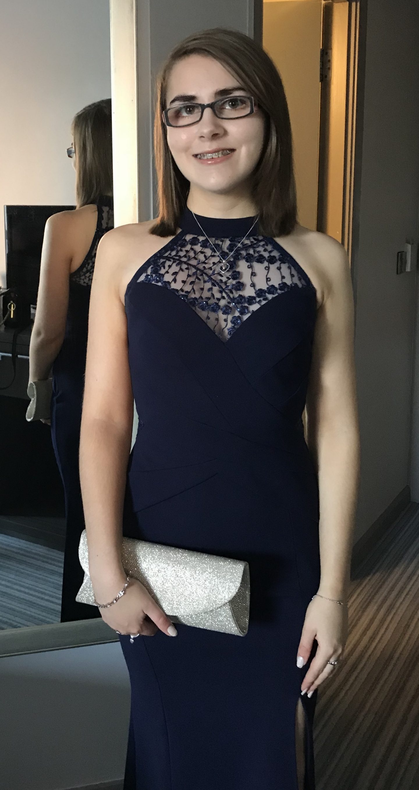A photo of Elin wearing a navy maxi dress which has a lace panel at the top extending into a high halter neck, there is also a small slit which goes up to just above her left knee. She wears it with a gold sparkly clutch bag.
