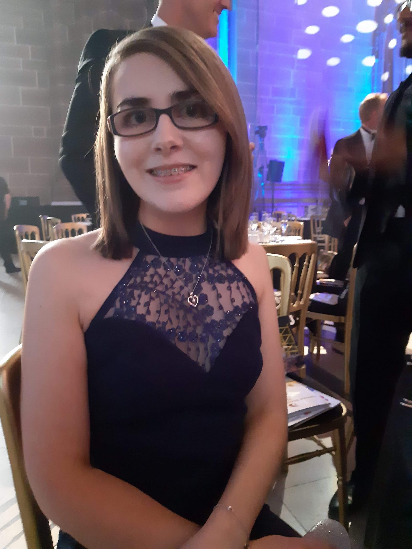 A photo of Elin sitting by a table at the national diversity awards, she wears a navy maxi dress which has a high halterneck and lace insert