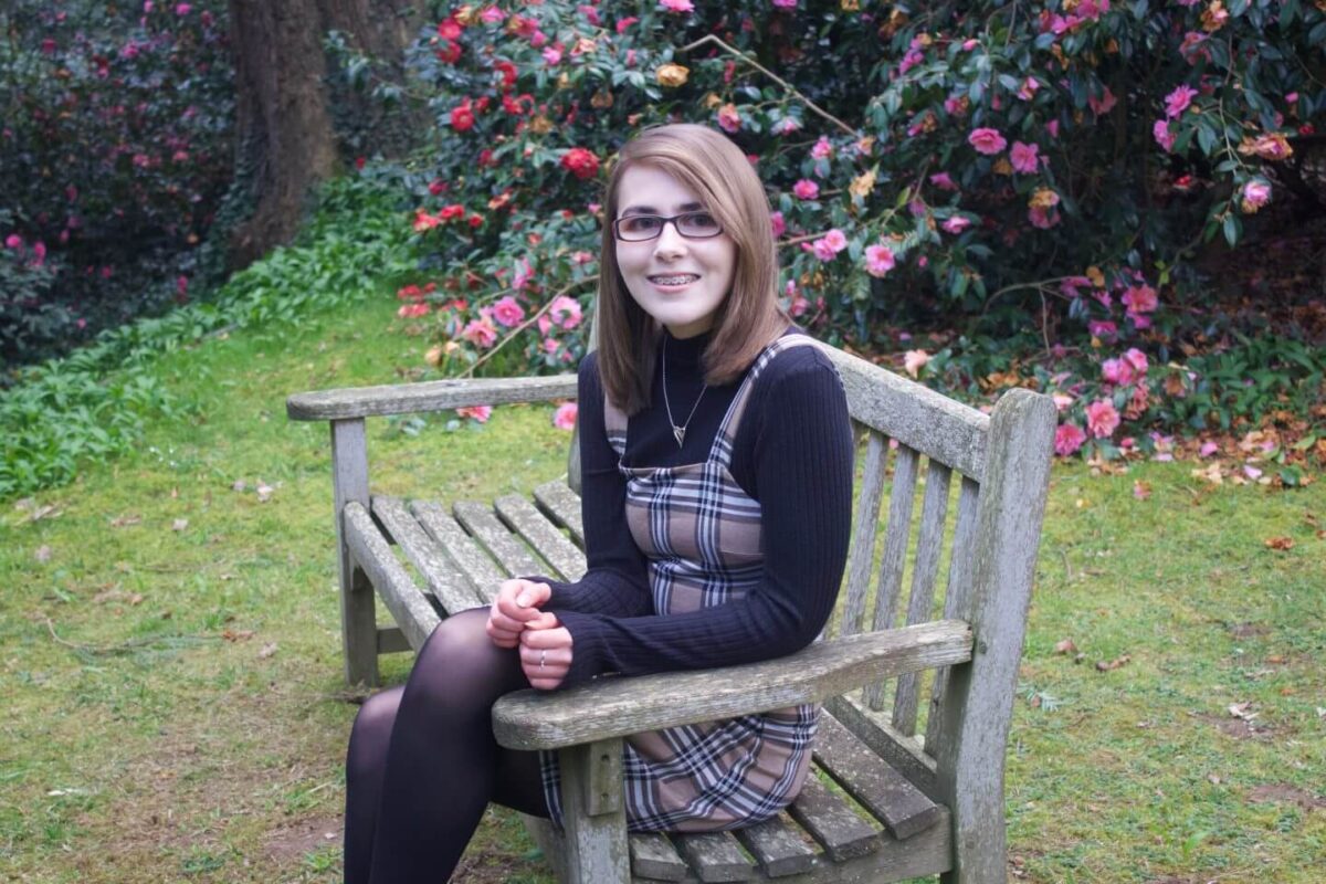 A photo of Elin sitting on a bench, there are pink flowers in the background