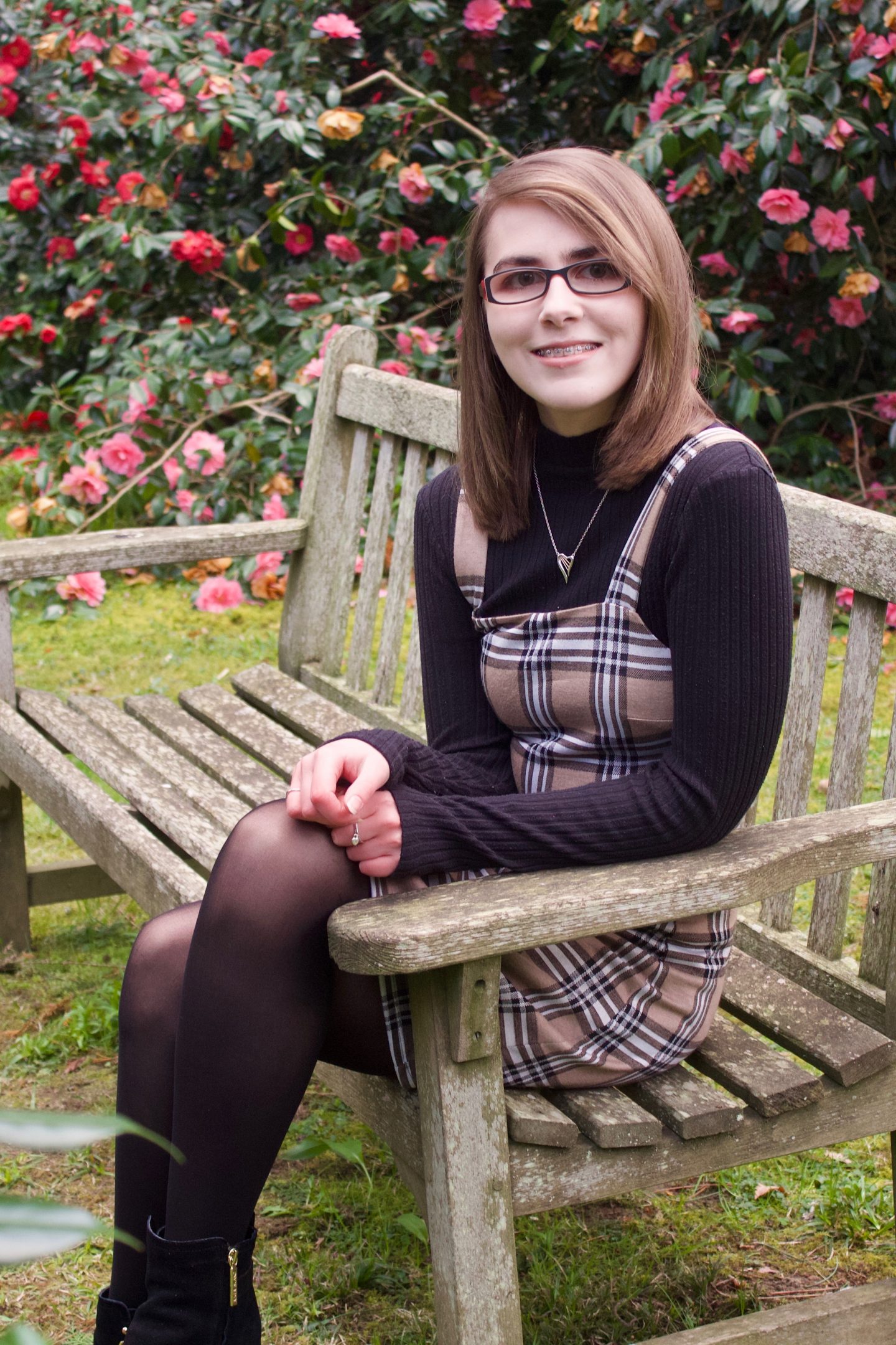 A photo of Elin sitting on a bench, she is wearing a beige check pinafore over a black high neck top