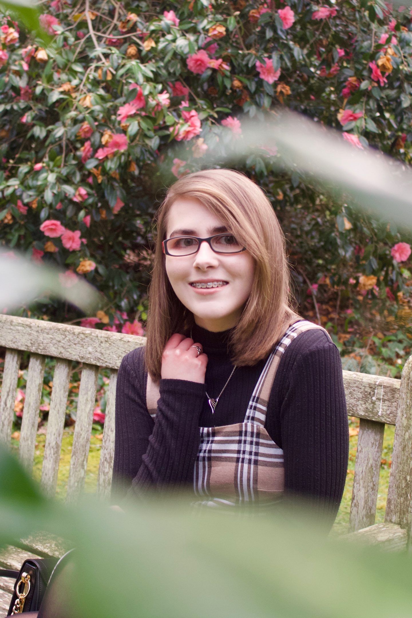 A photo of Elin sitting on a bench, she is framed by some green leaves which are out of focus