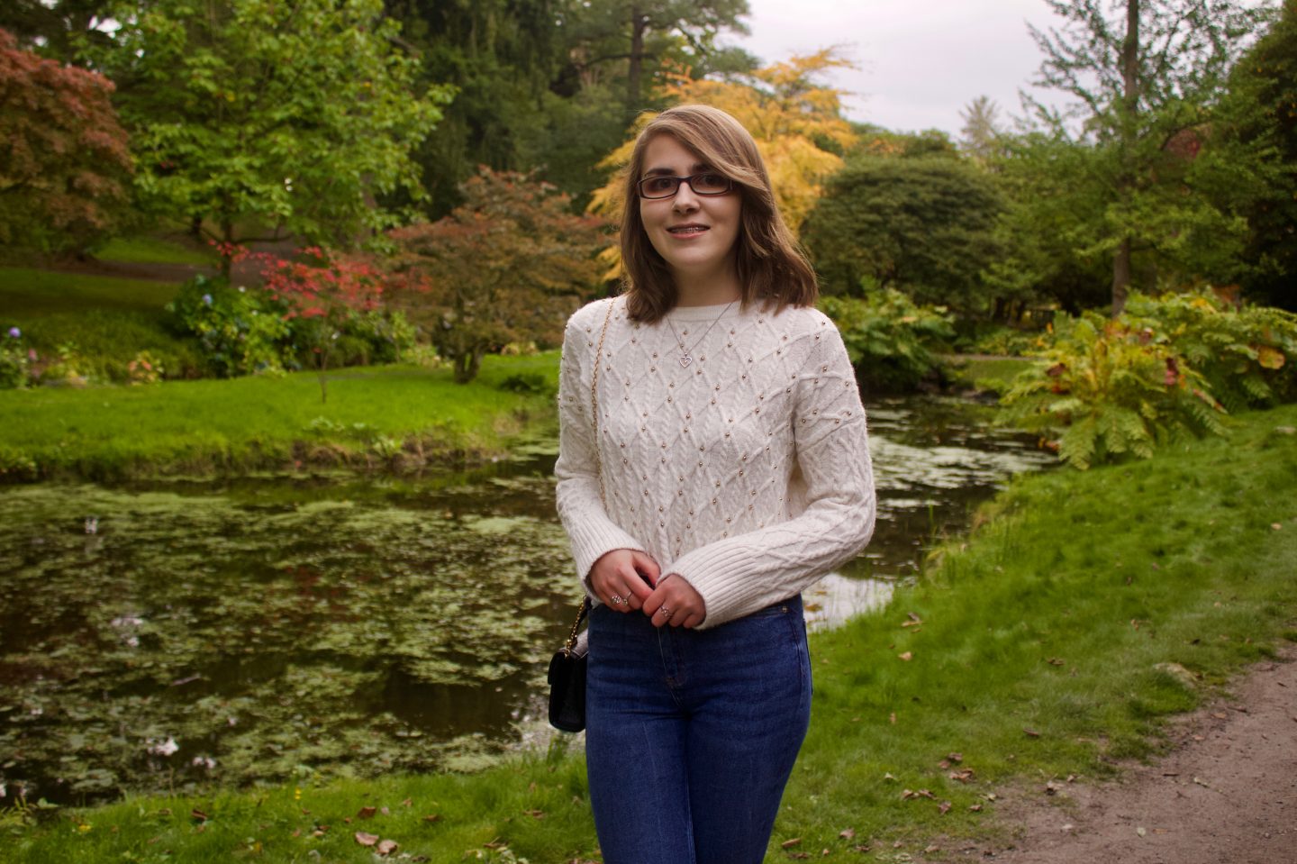 A photo of Elin standing in front of a small lake with trees and flowers in the background, she is smiling at the camera and wears a cream knitted crop jumper which has small gold studs on it and blue skinny jeans