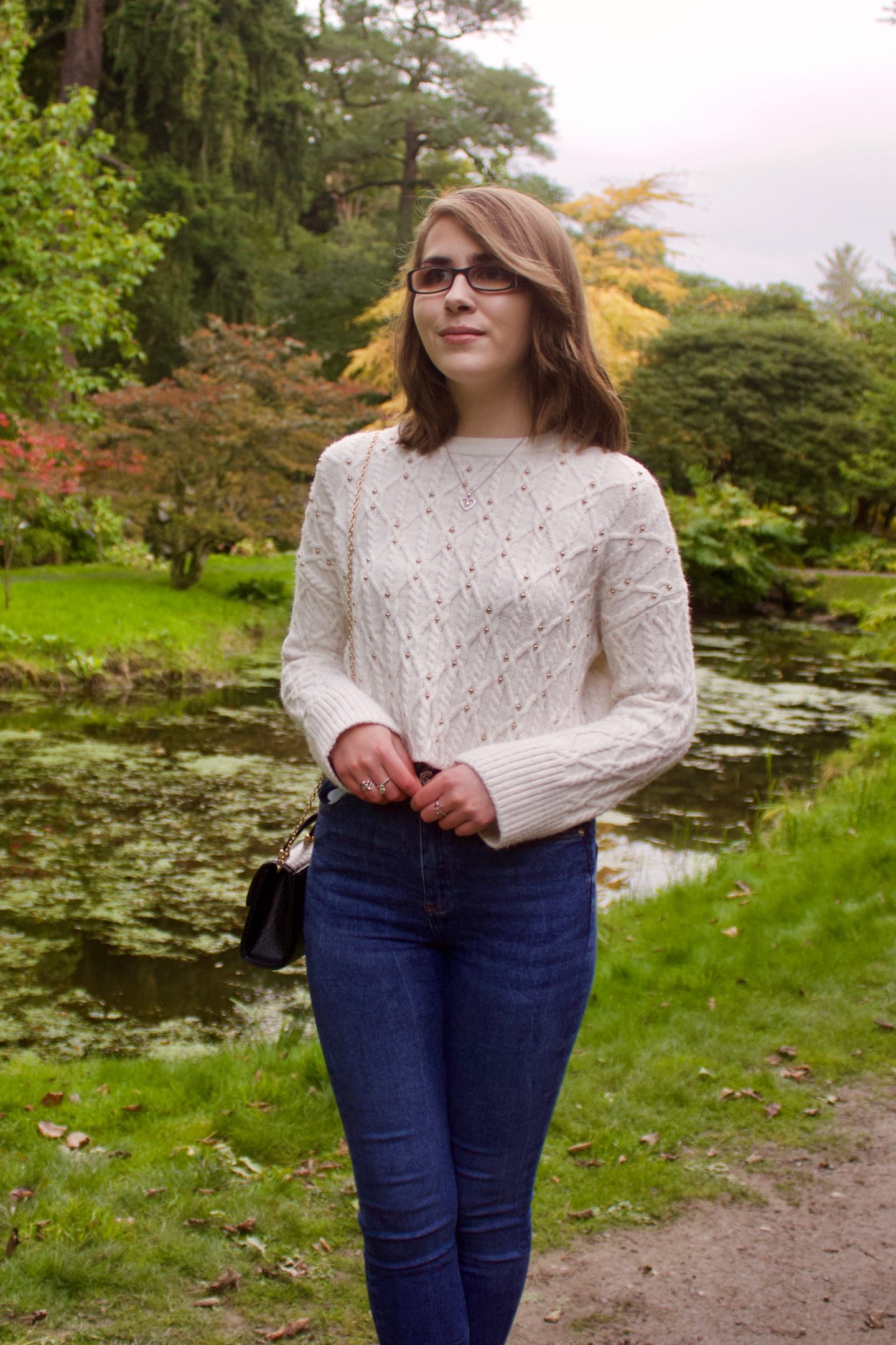 A photo of Elin standing in front of a small lake, the trees in the background reflect on the water and she is looking away from the camera