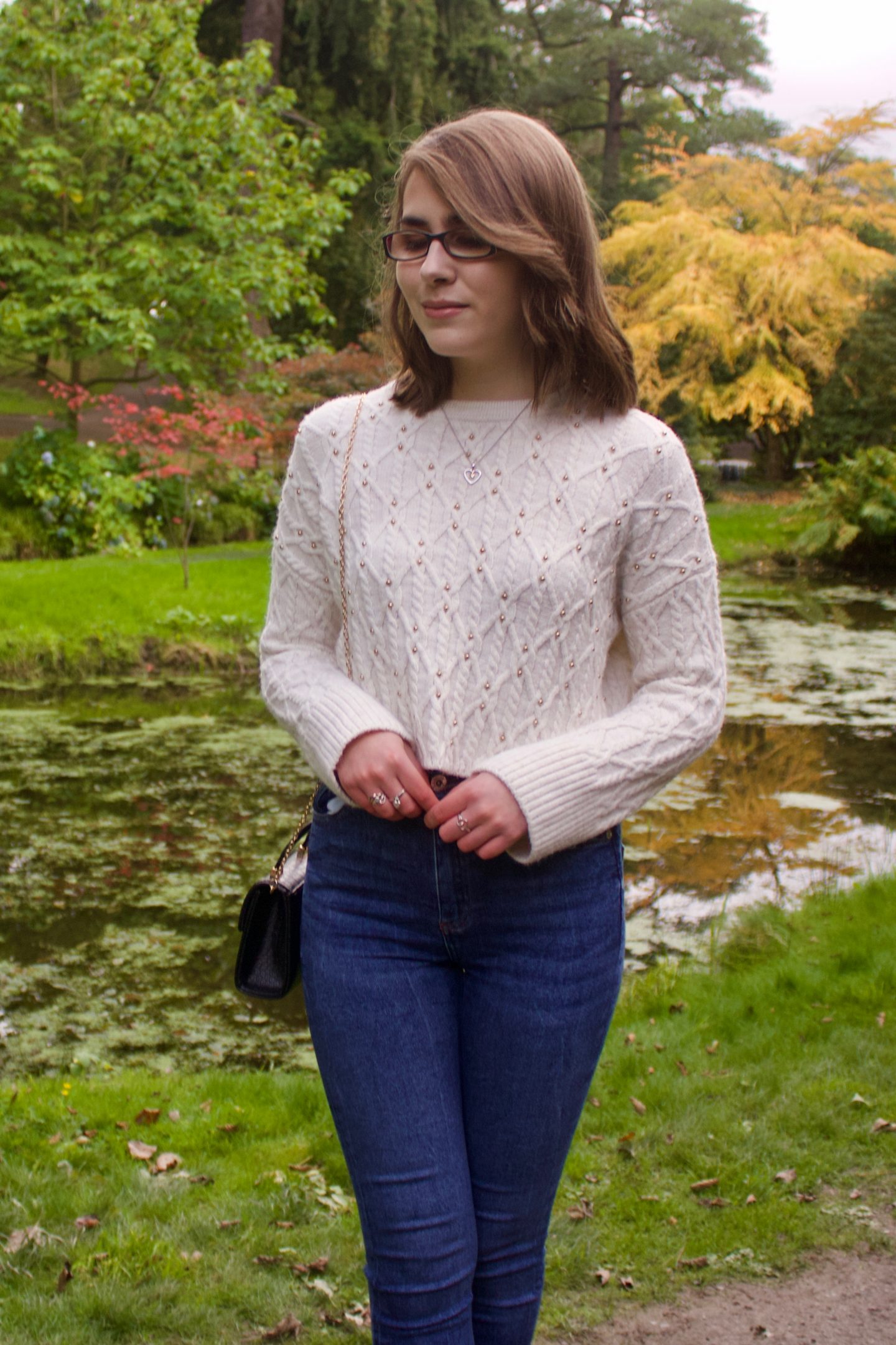 A photo of Elin standing in front of a small lake, she is looking down and there are trees in the background, one has yellow leaves on it