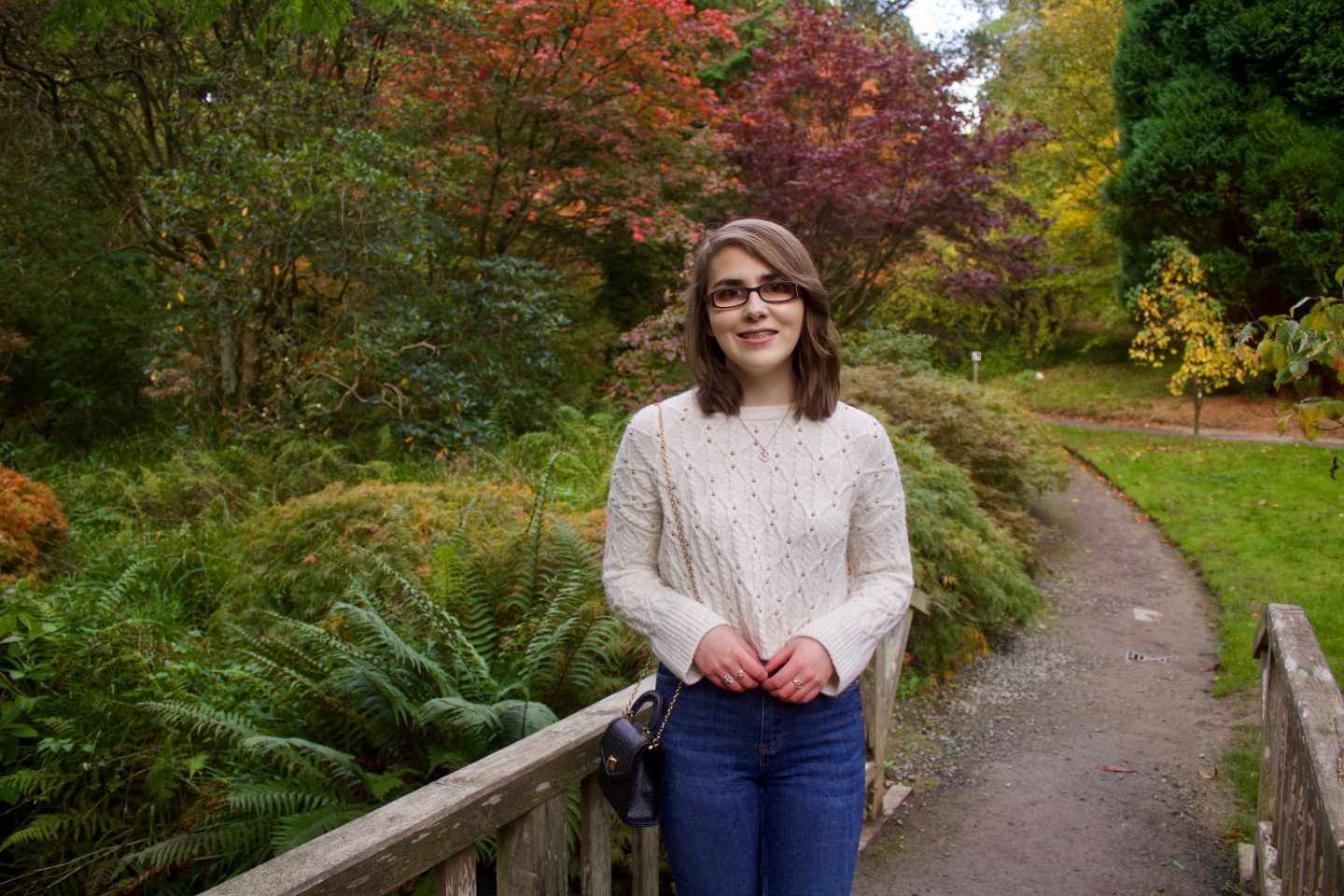 A photo of Elin standing on a small wooden bridge, there are lots of colourful trees and plants behind her, one of the trees has red leaves on it