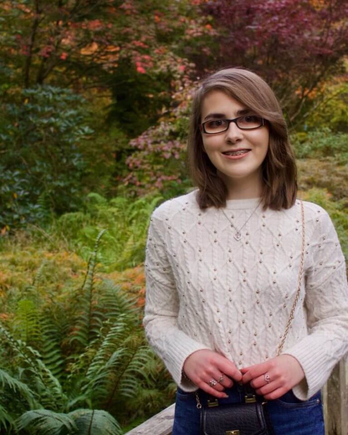 A photo of Elin standing on a small wooden bridge, there are lots of colourful trees and plants behind her, one of the trees has red leaves on it