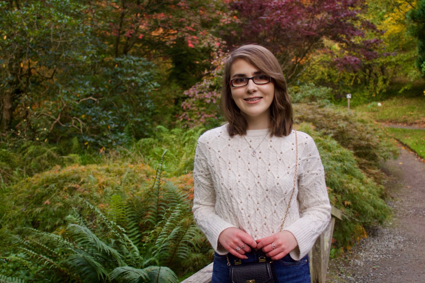 A photo of Elin standing on a small wooden bridge, there are lots of colourful trees and plants behind her, one of the trees has red leaves on it