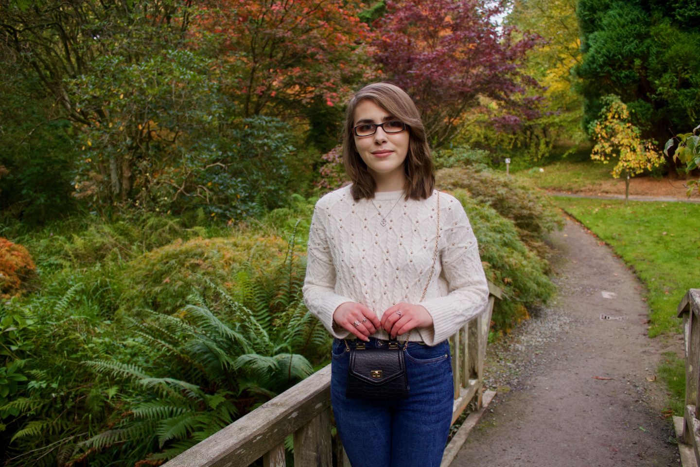 A photo of Elin leaning against a wooden bridge, she wears a cropped cream knitted jumper with skinny belted blue jeans and a mini black cross body bag which has a gold chain. There are colourful trees and plants in the background - Let's talk about self-confidence
