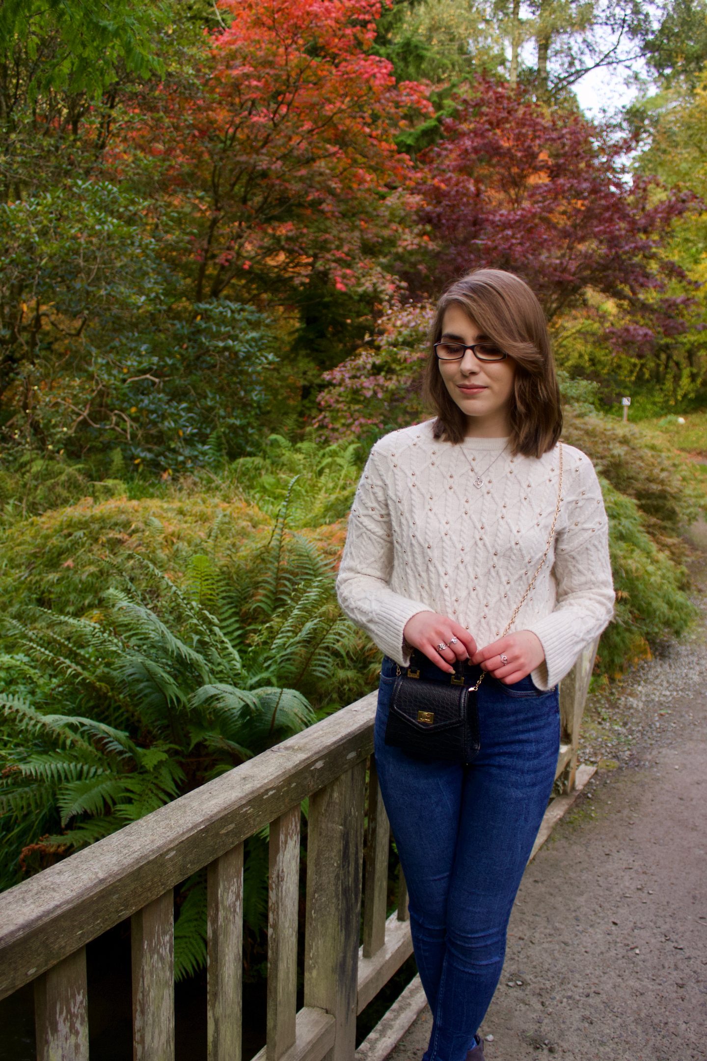 A photo of Elin standing on a wooden bridge, she is looking down and she wears a cream knitted jumper, blue skinny jeans and a black cross body bag