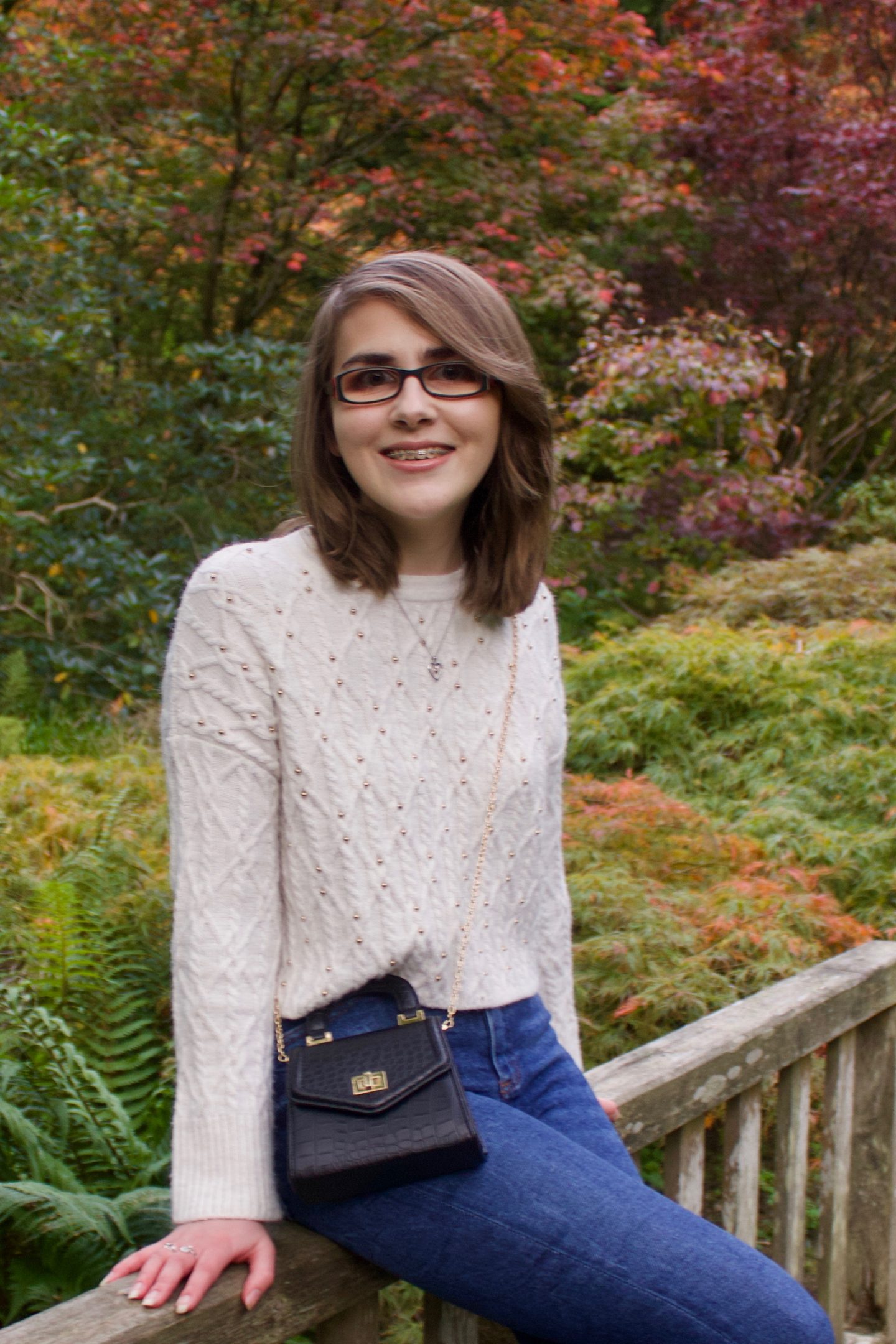 A photo of Elin sitting on a small wooden bridge, she is smiling at the camera and there are pretty trees and flowers in the background, all in pretty autumnal colours