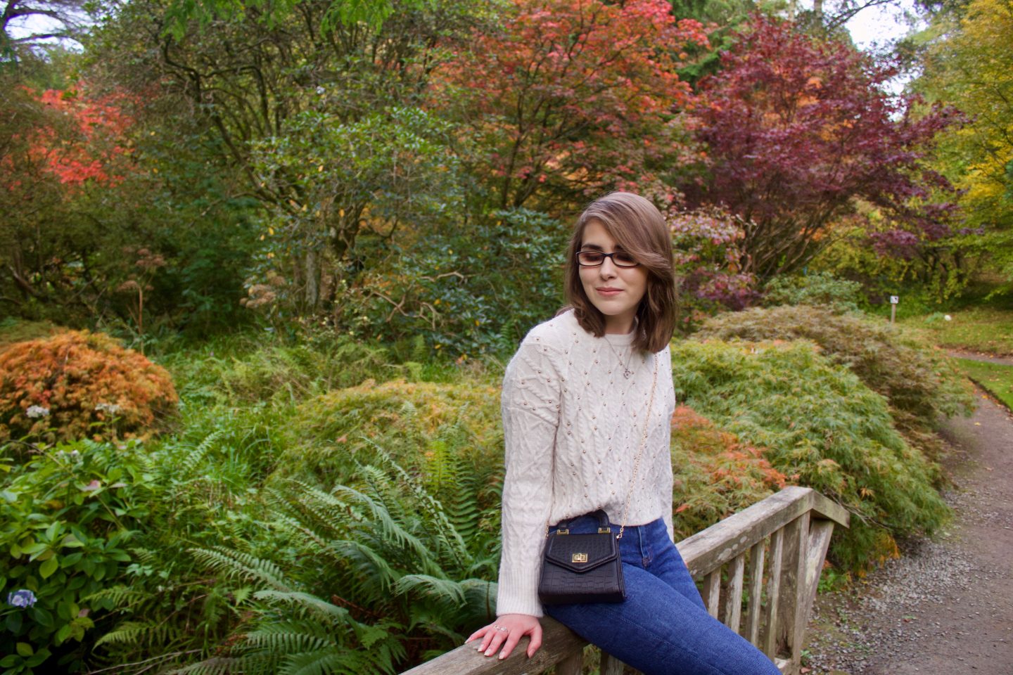 A photo of Elin sitting on a small wooden bridge, there are colourful trees and flowers in the background, all in pretty autumn colours. Elin is looking down
