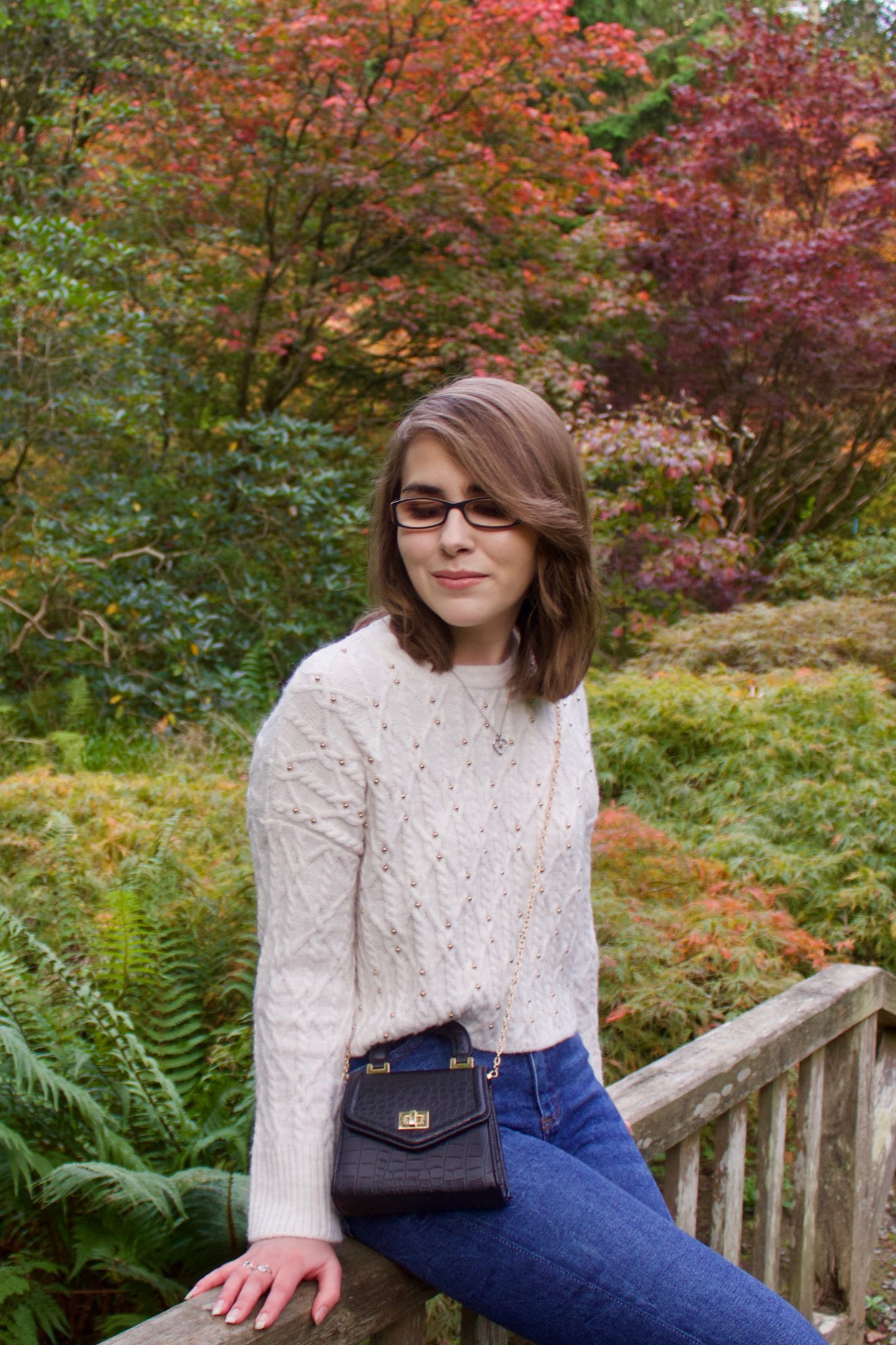 A photo of Elin sitting on a small wooden bridge, there are colourful trees and flowers in the background, all in pretty autumn colours. Elin is looking down