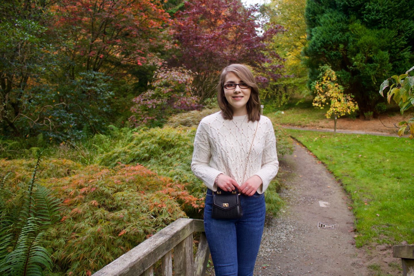 A photo of Elin standing in front of trees which have colourful autumnal leaves, she wears a cream knitted jumper which has dainty gold embelished studs on it, a pair of blue skinny jeans and a small black cross body bag which has a gold chain