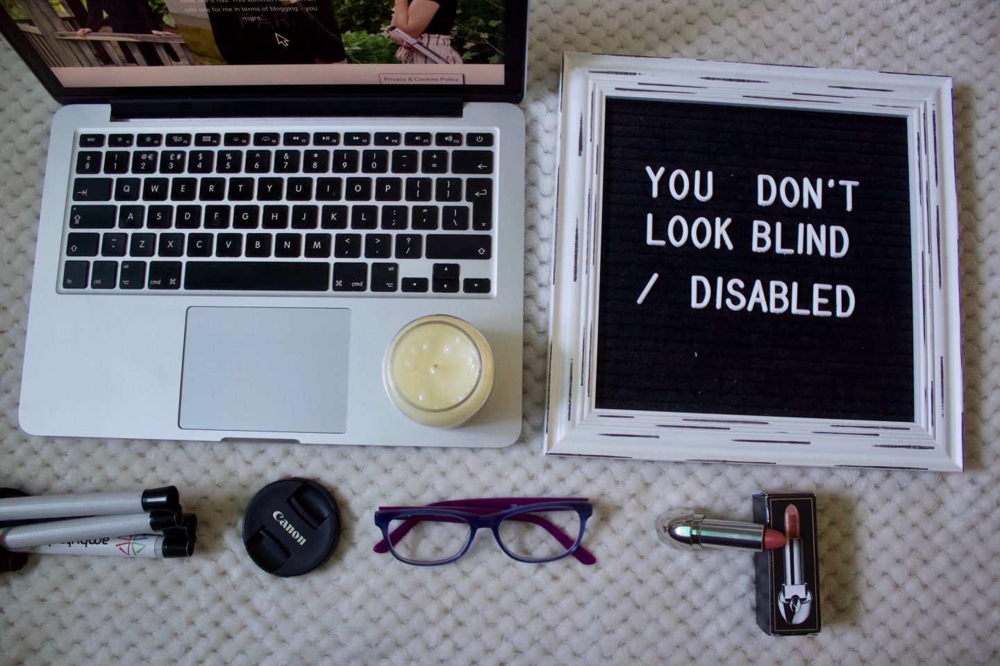 A photo of a black felt letter board which has a thick white frame, the words "You don't look blind/disabled" are on the board in white letters. A macbook is beside it with a candle on the keyboard