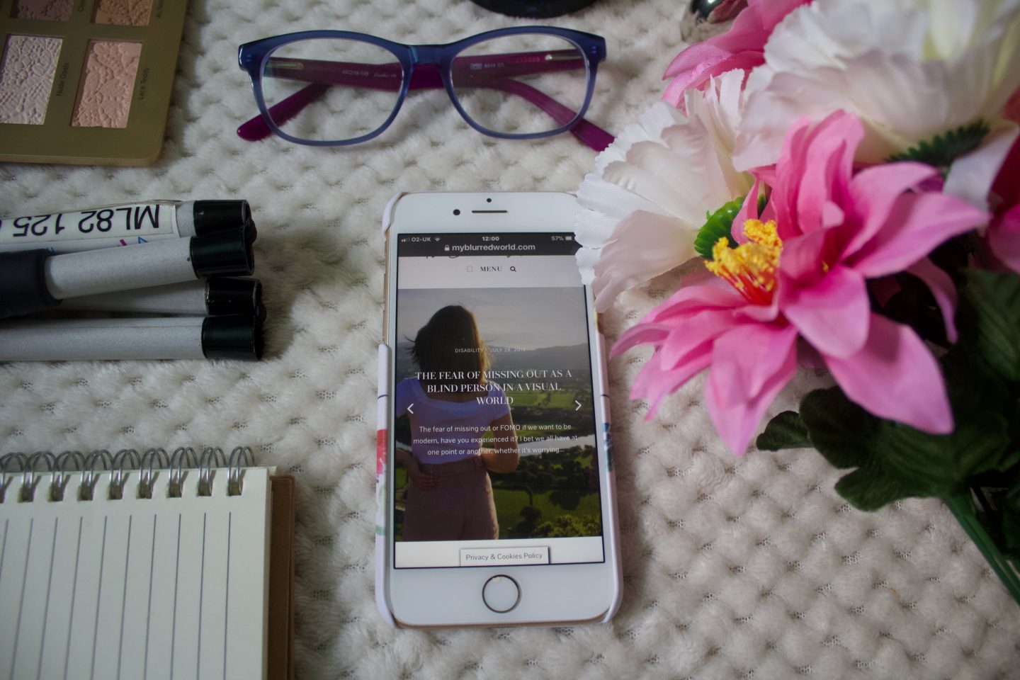 A photo of my blog's homepage on my phone, there are pink artificial flowers beside it and a pair of glasses