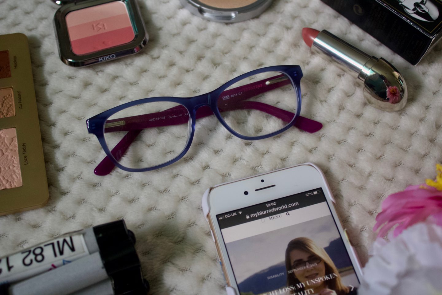 A photo of dark blue framed glasses, there are a few makeup products around them such as a lipstick and a blush